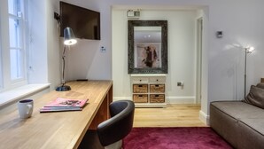 A room with a wooden desk, a black chair, a red carpet, a grey sofa, a TV mounted on the wall, and a mirror with a decorative frame. - Eson2 - The York Street Nest (London)
