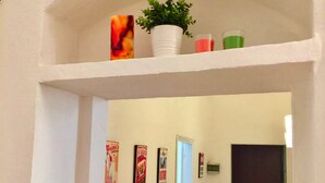 Um corredor com um arco branco que dá para um quarto. Há três cartazes emoldurados na parede, um vaso de planta e uma pequena prateleira com um vaso e uma vela.