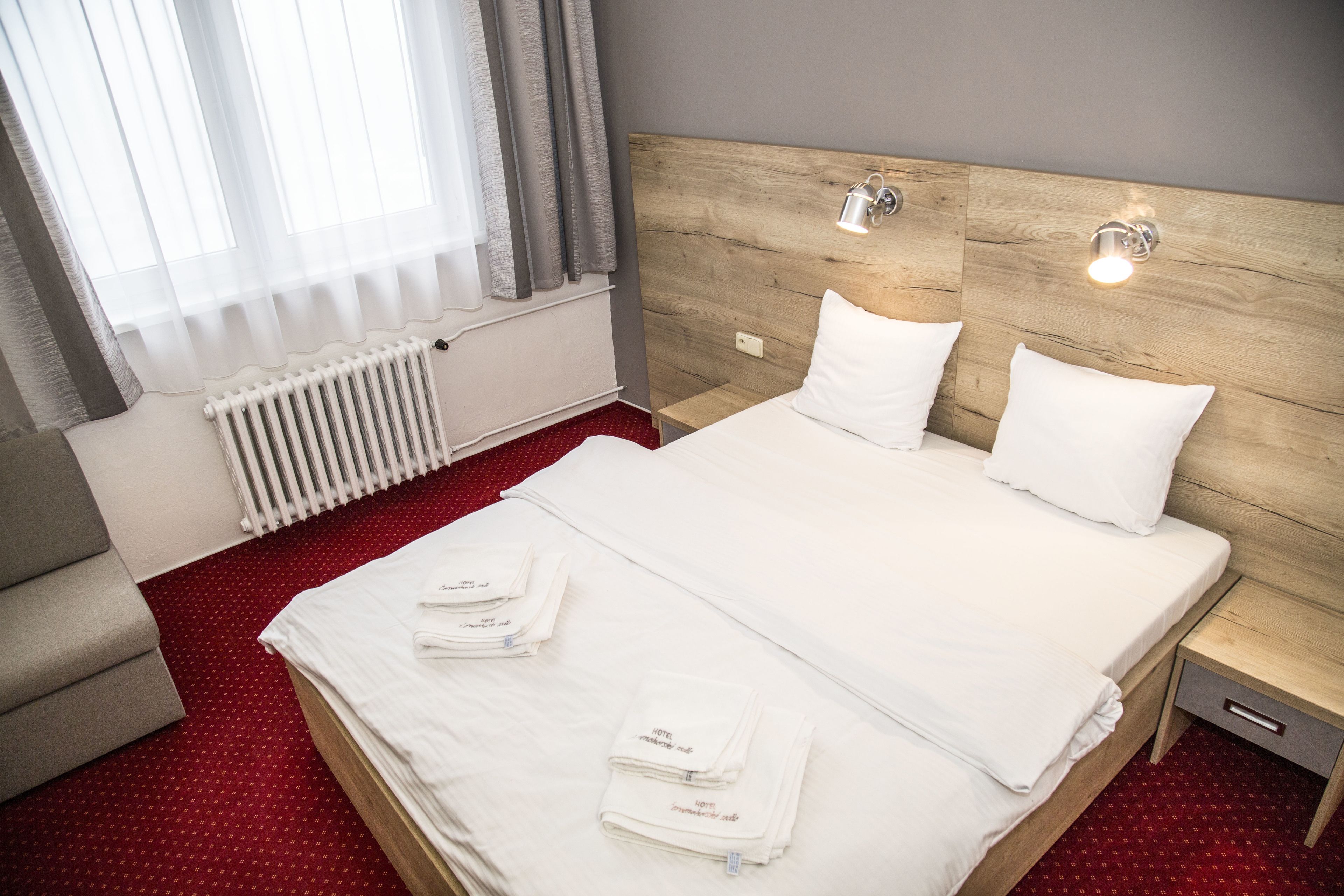 A hotel room with a large bed, two bedside tables, a nightstand, a radiator, and a window with curtains.