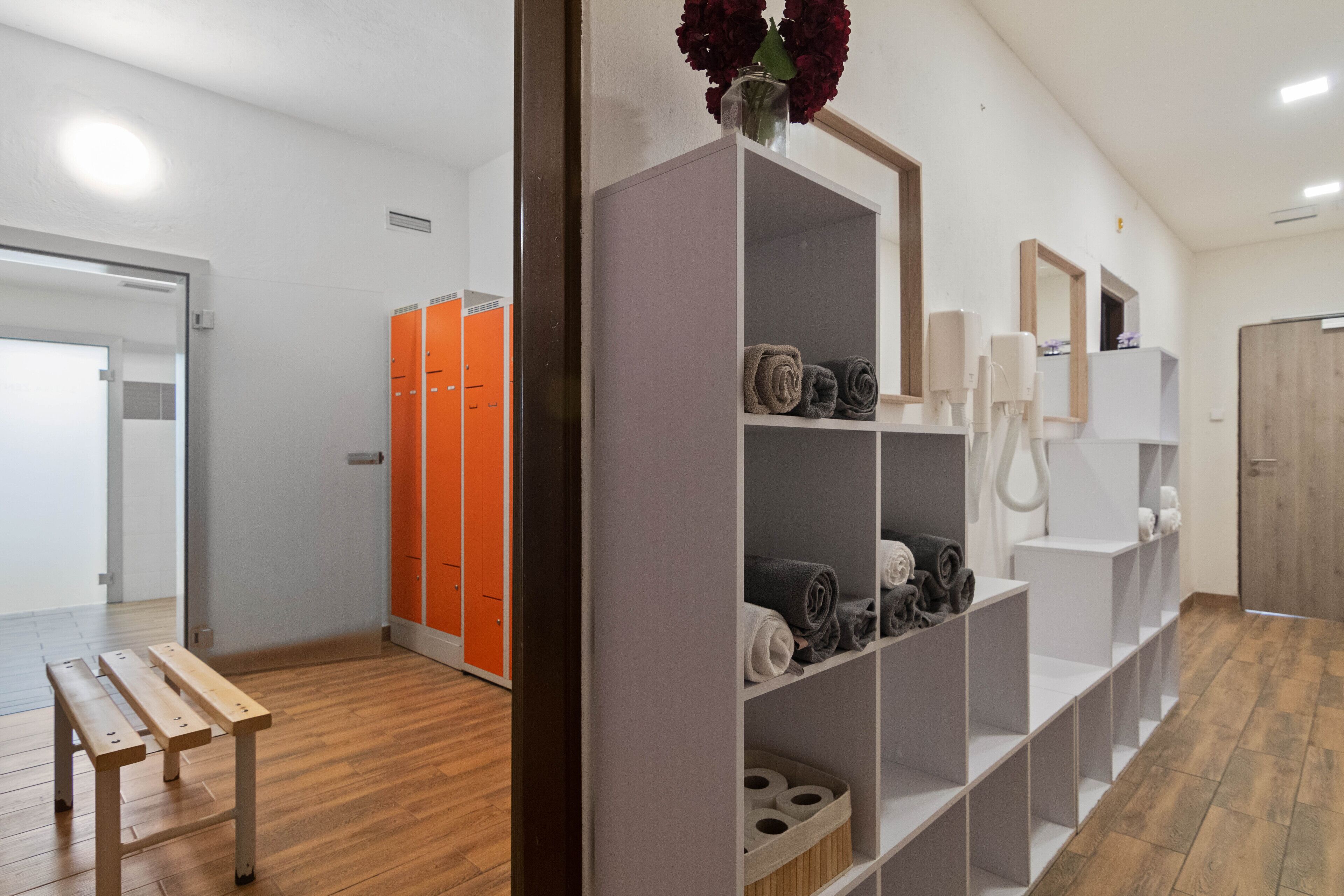 A hotel room with a wooden floor, a bench, a wardrobe with shelves, and orange lockers in the background.