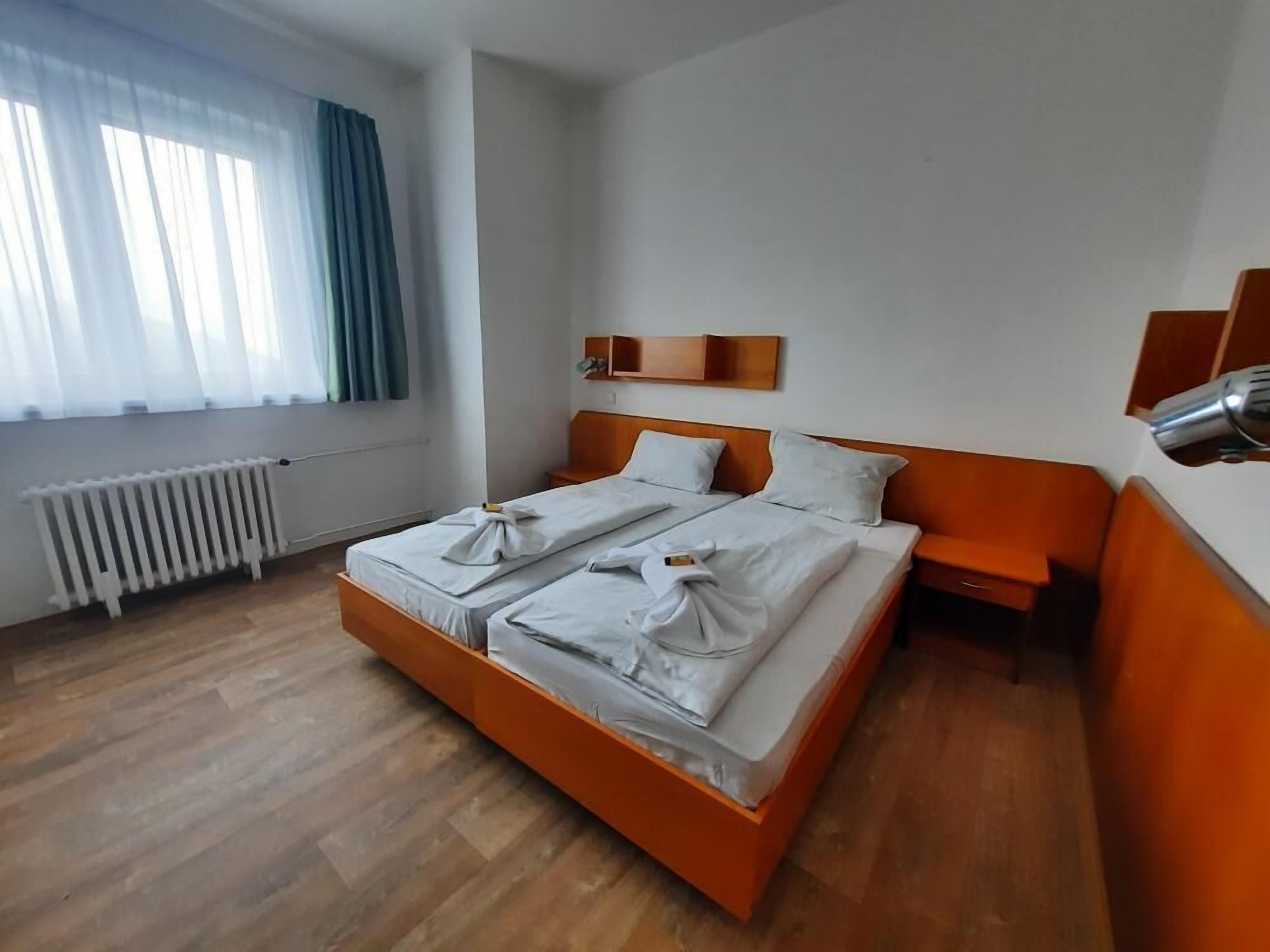 A double bed with white linens, a wooden headboard, a bedside table, a small shelf, and a radiator.