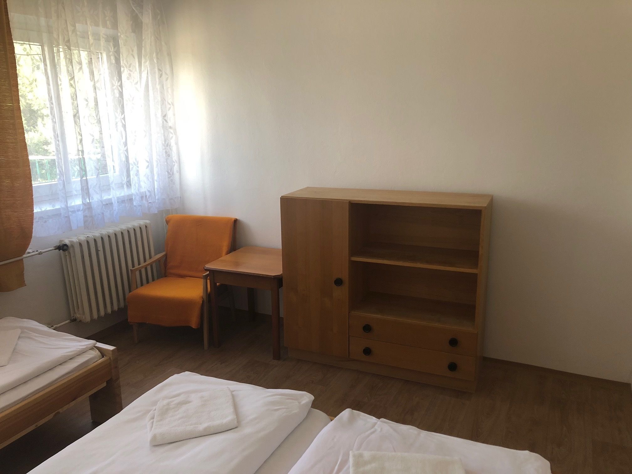 A room with two beds, a wooden cabinet with drawers, an orange chair, and a window with sheer curtains.