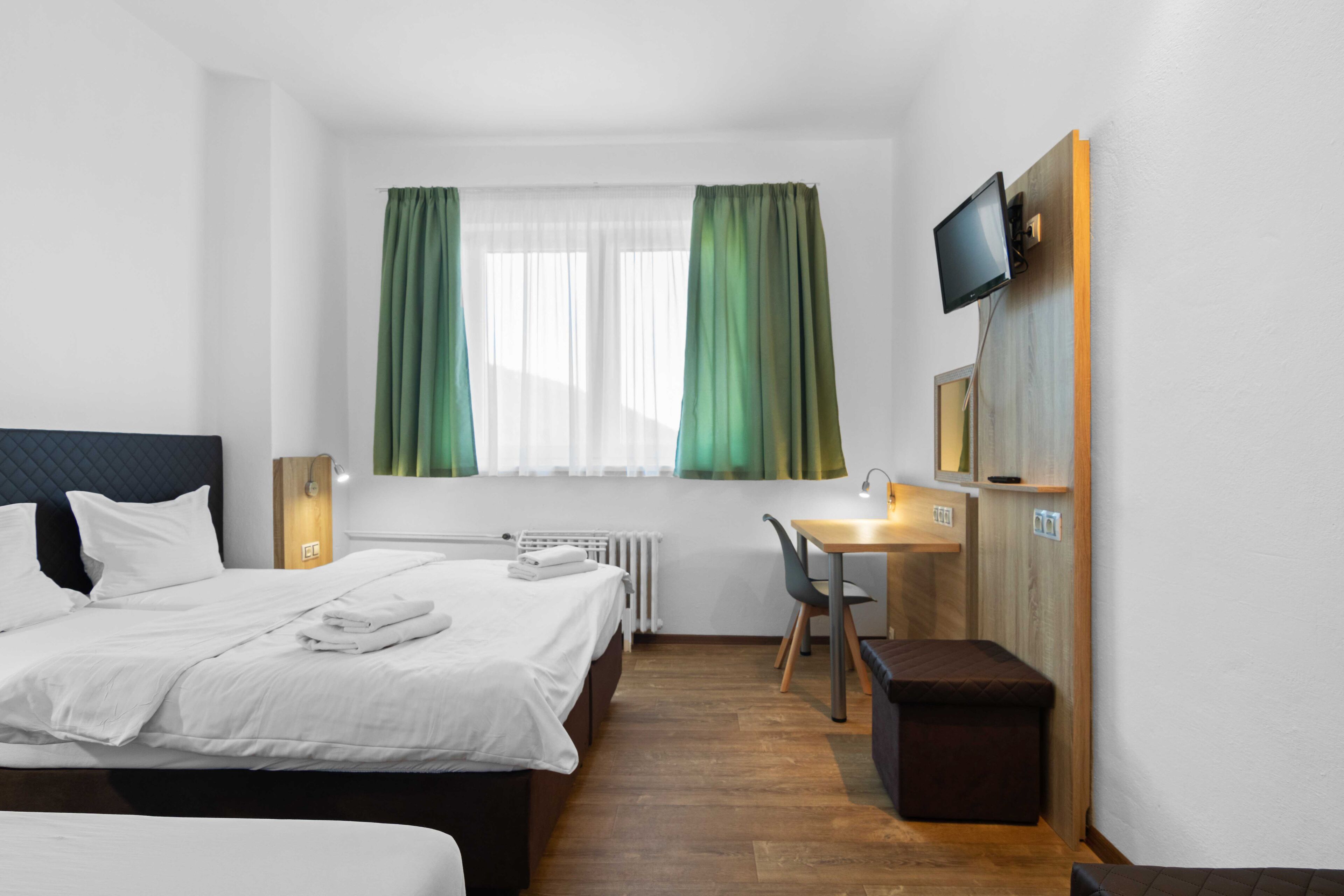 A hotel room with a bed, a desk, a chair, a TV mounted on the wall, and a window with curtains.