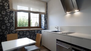 Une cuisine moderne avec un plan de travail blanc, des appareils en acier inoxydable, une fenêtre ornée d’un rideau à motifs floraux et un coin repas avec une table et des chaises.