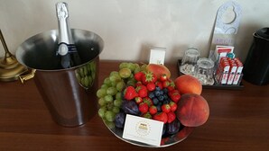 Un frutero con uvas, fresas y melocotones, una botella de champán y una bandeja con bombones Kinder y una copa.