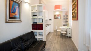 Una sala de estar moderna con un sofá de cuero negro, un estante blanco con libros y objetos decorativos, y un comedor con mesa y sillas.