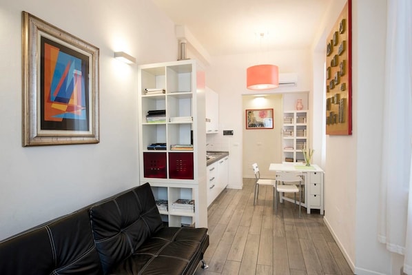 A modern living room with a black leather sofa, white shelving units, and a dining area with a table and chairs.