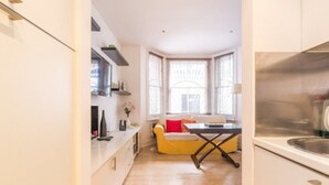 Une cuisine compacte avec des meubles blancs, un coin repas comprenant une chaise jaune et une table, et une fenêtre avec des stores.