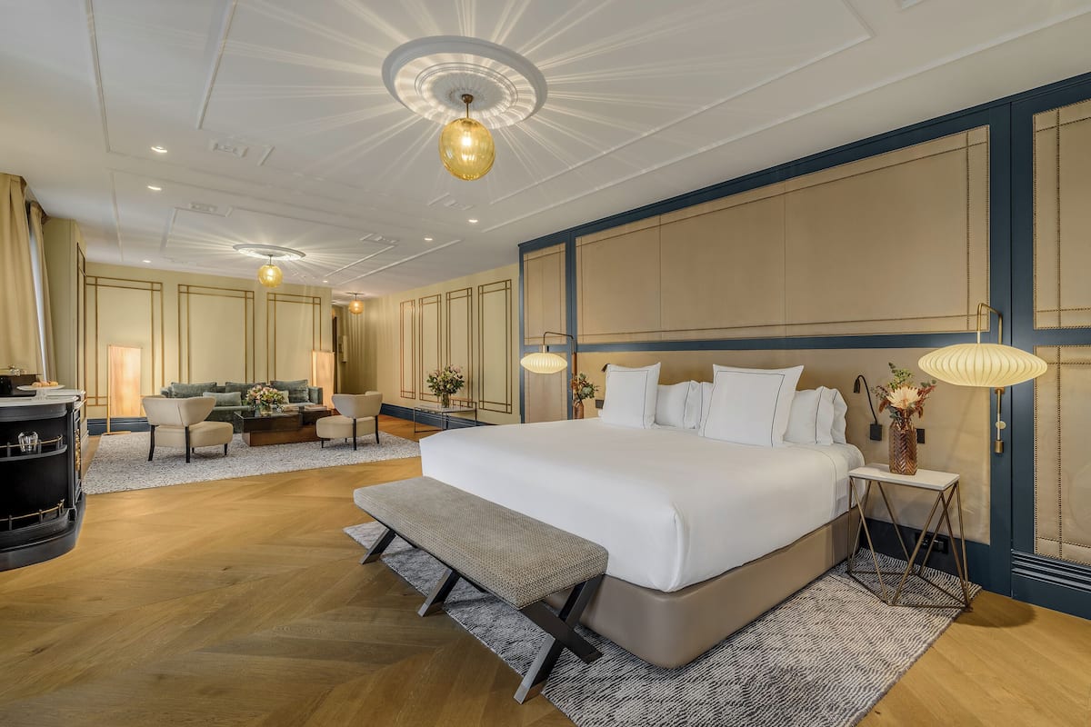 Une chambre spacieuse avec un grand lit, un banc, un bureau et un coin salon comprenant un canapé et des fauteuils.