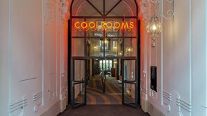 An ornate hallway with a chandelier, a neon sign reading "COOL ROOMS", and a staircase leading to an upper level.