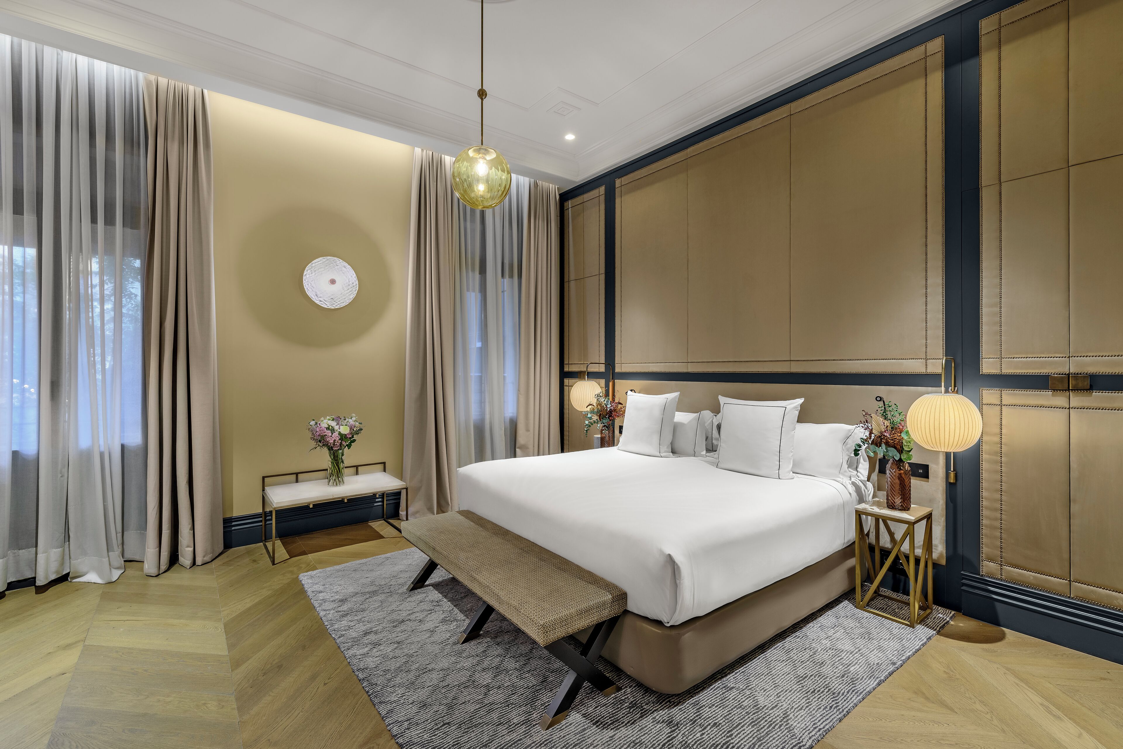 Una habitación de hotel moderna con una cama grande, un banco, una mesita de noche, una lámpara y vistas al exterior a través de cortinas translúcidas.