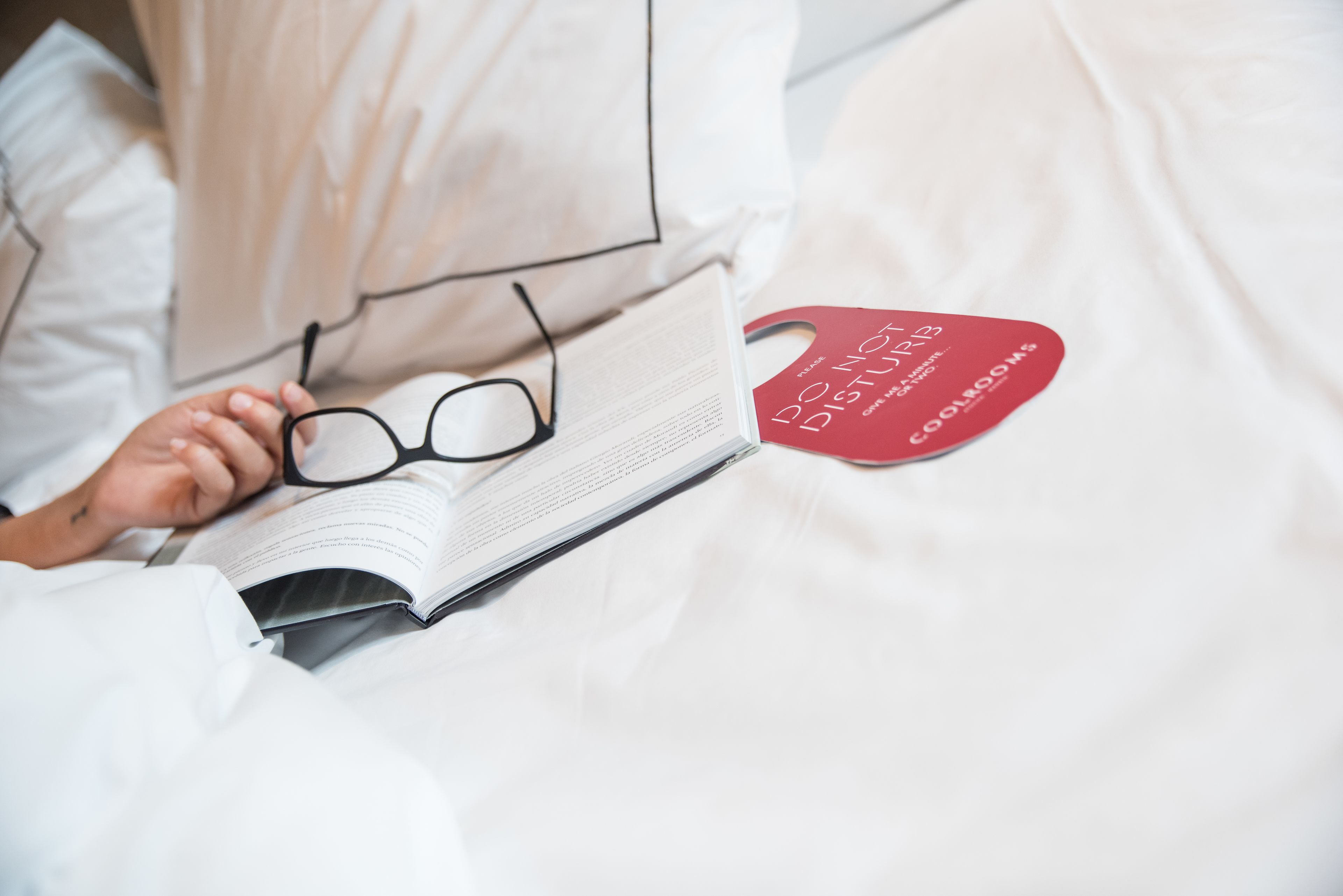 A person reading a book on a bed with white bedding and a red bookmark.