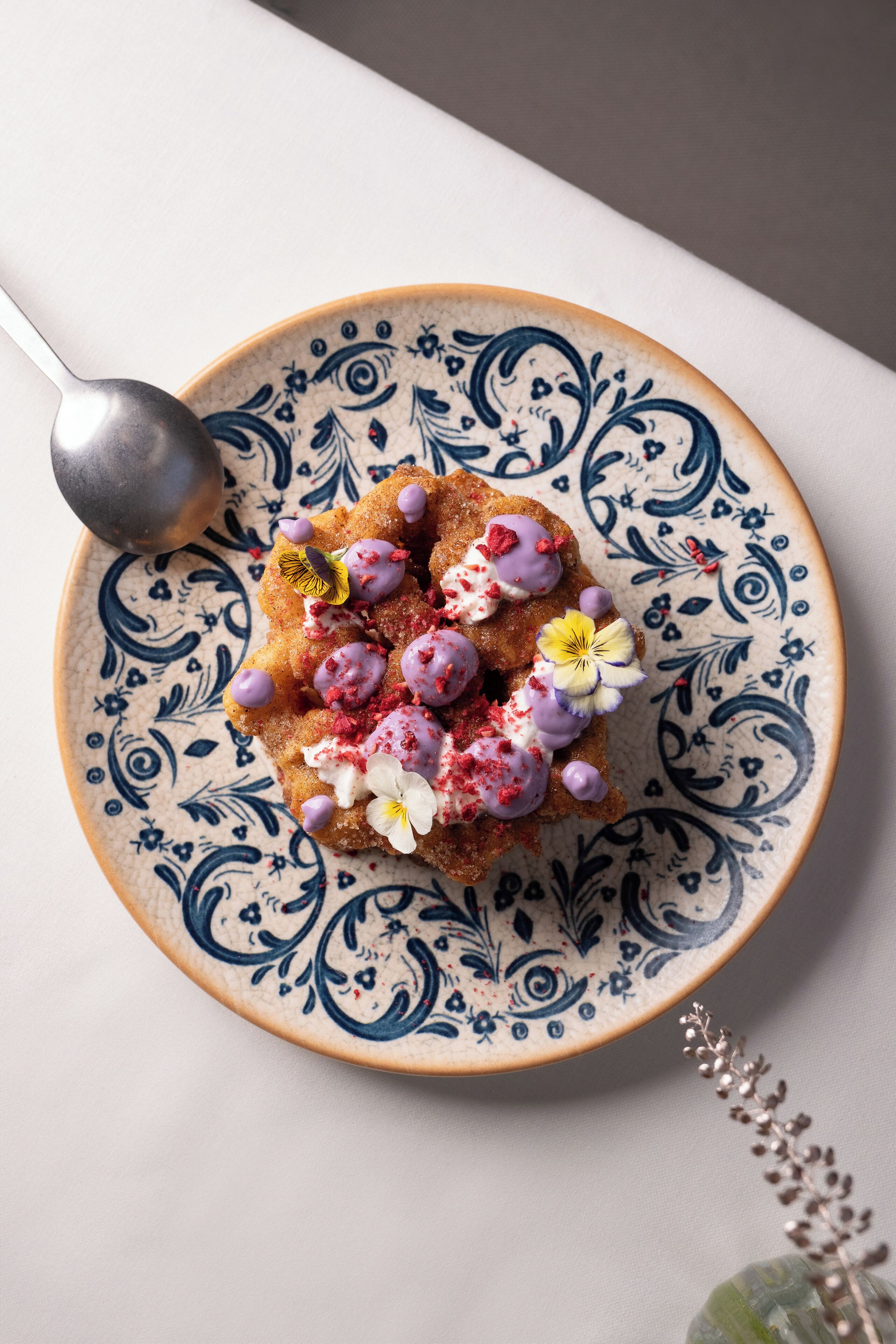 A plate with a pastry topped with berries and flowers, a spoon, and a decorative vase.
