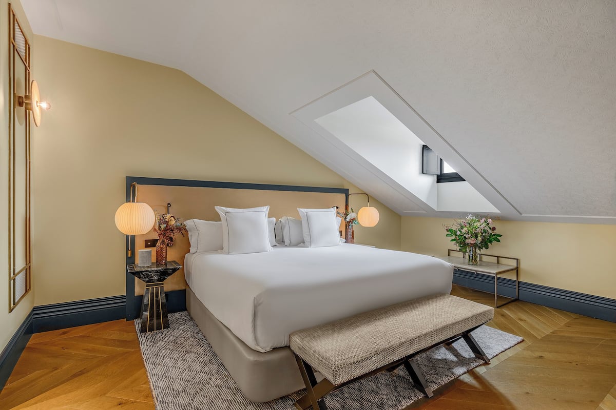 Une chambre avec un grand lit, une lucarne, une table de chevet avec une lampe et un banc.