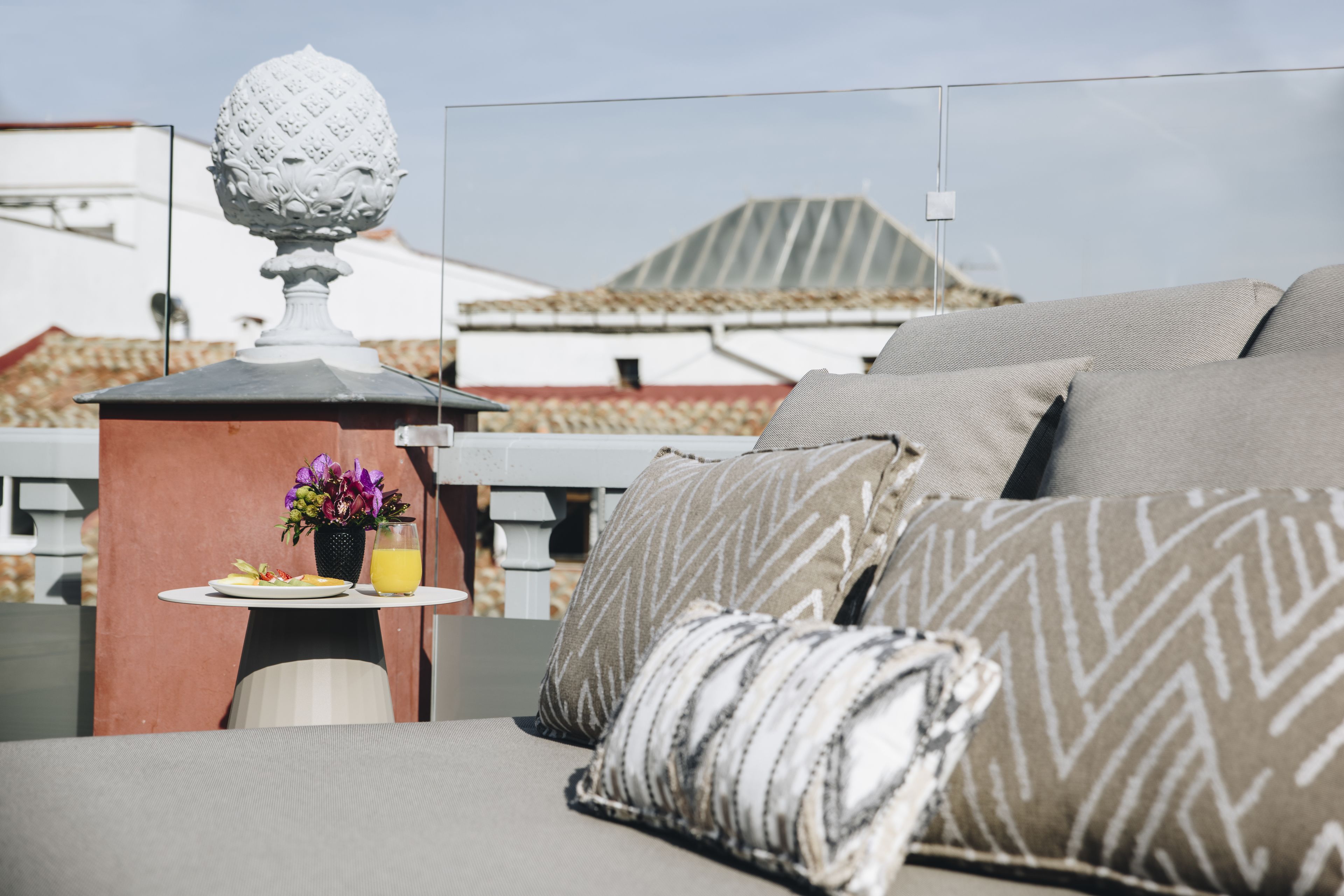 A rooftop terrace with a sofa, pillows, a table with a drink and flowers, and a decorative globe.