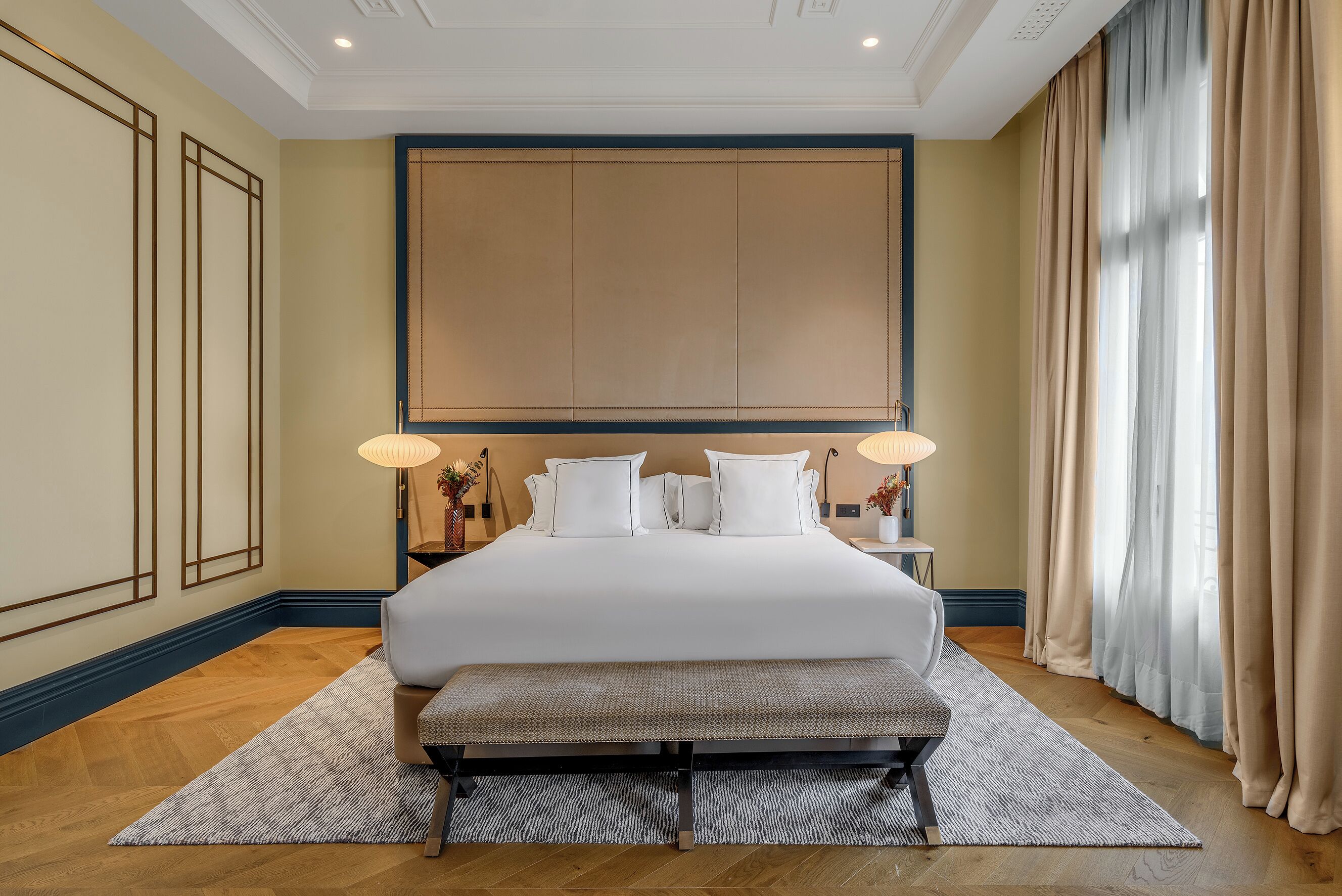 A large bed with white linens, a bench, and two bedside lamps in a hotel room with wooden flooring and beige walls.