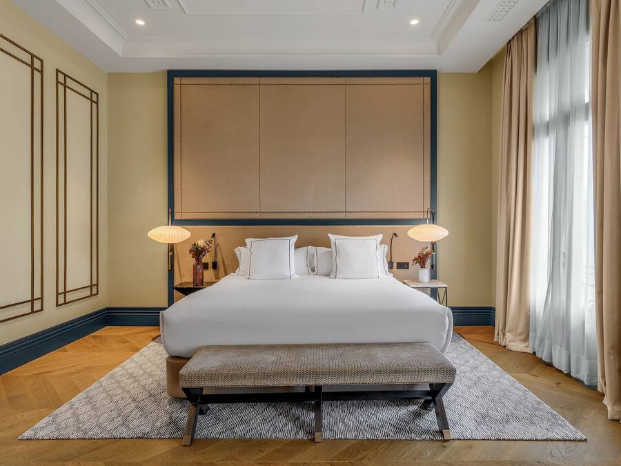 A large bed with white linens, a bench, and two bedside lamps in a hotel room with wooden flooring and beige walls.