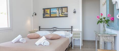 A neatly made bed with pillows and rolled towels, a bedside table with a vase of flowers, and a painting on the wall.