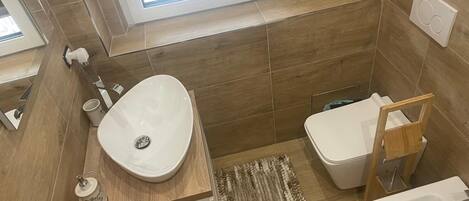 Un baño moderno con un ventanal grande, un tocador de madera, un lavamanos, un inodoro y una alfombra pequeña.