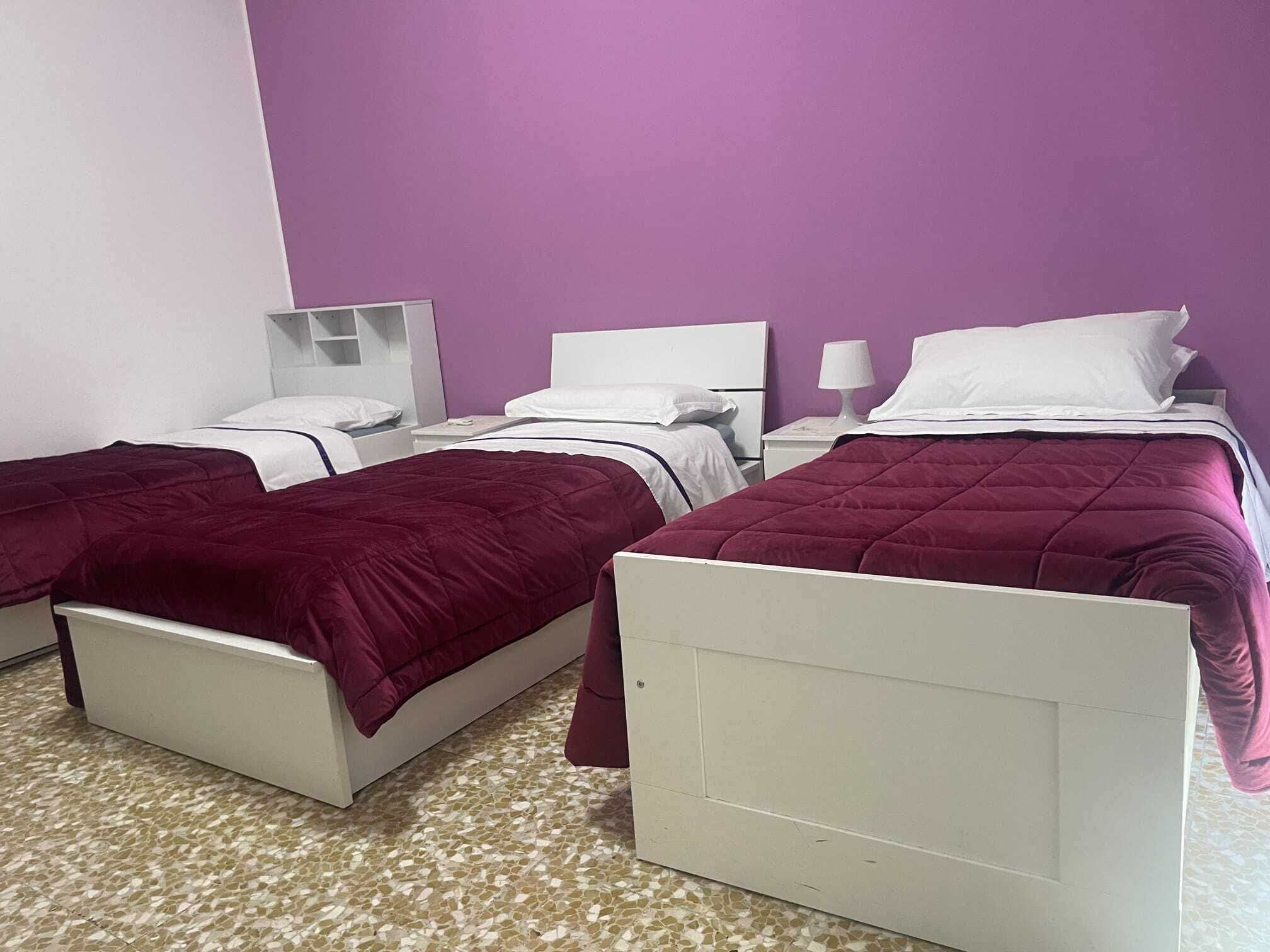Camera d'albergo con due letti, ognuno con testata bianca e copripiumino viola. Su ogni letto, un comodino con una lampada.
