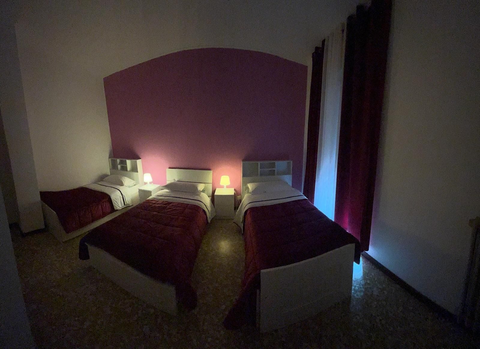 A hotel room with two beds, each with a white headboard and red bedding. There are bedside tables with lamps, and the room has a purple accent wall.