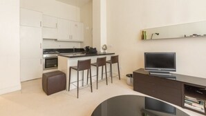 Una cucina moderna con mobili bianchi, un'isola centrale con sgabelli da bar, forno integrato e televisore a schermo piatto su un supporto in legno scuro.