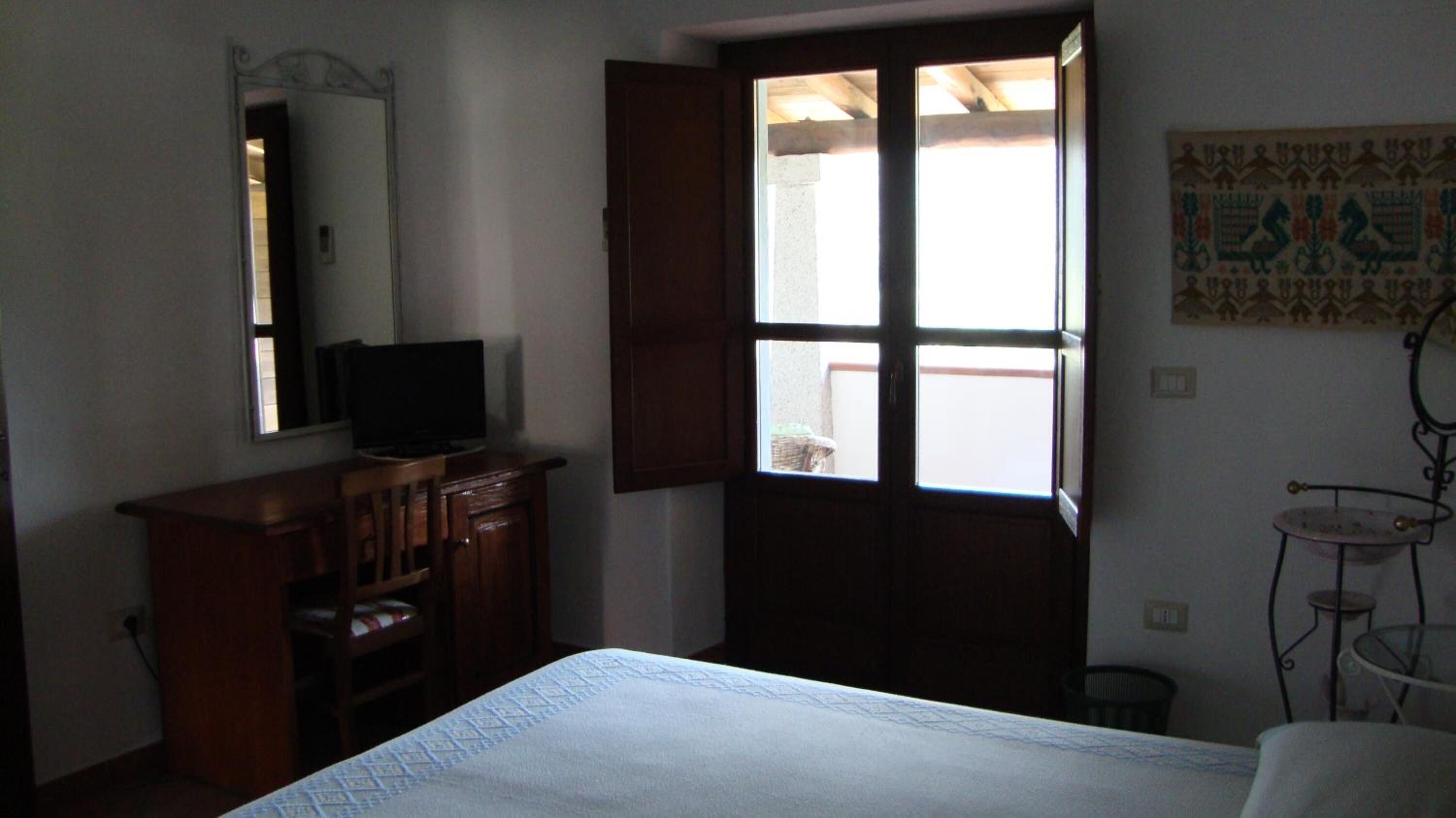 Ein Schlafzimmer mit einem Bett, einer Holzkommode, einem Fernseher, einem Fenster mit geöffneten Lamellen und einem an der Wand befestigten Spiegel.