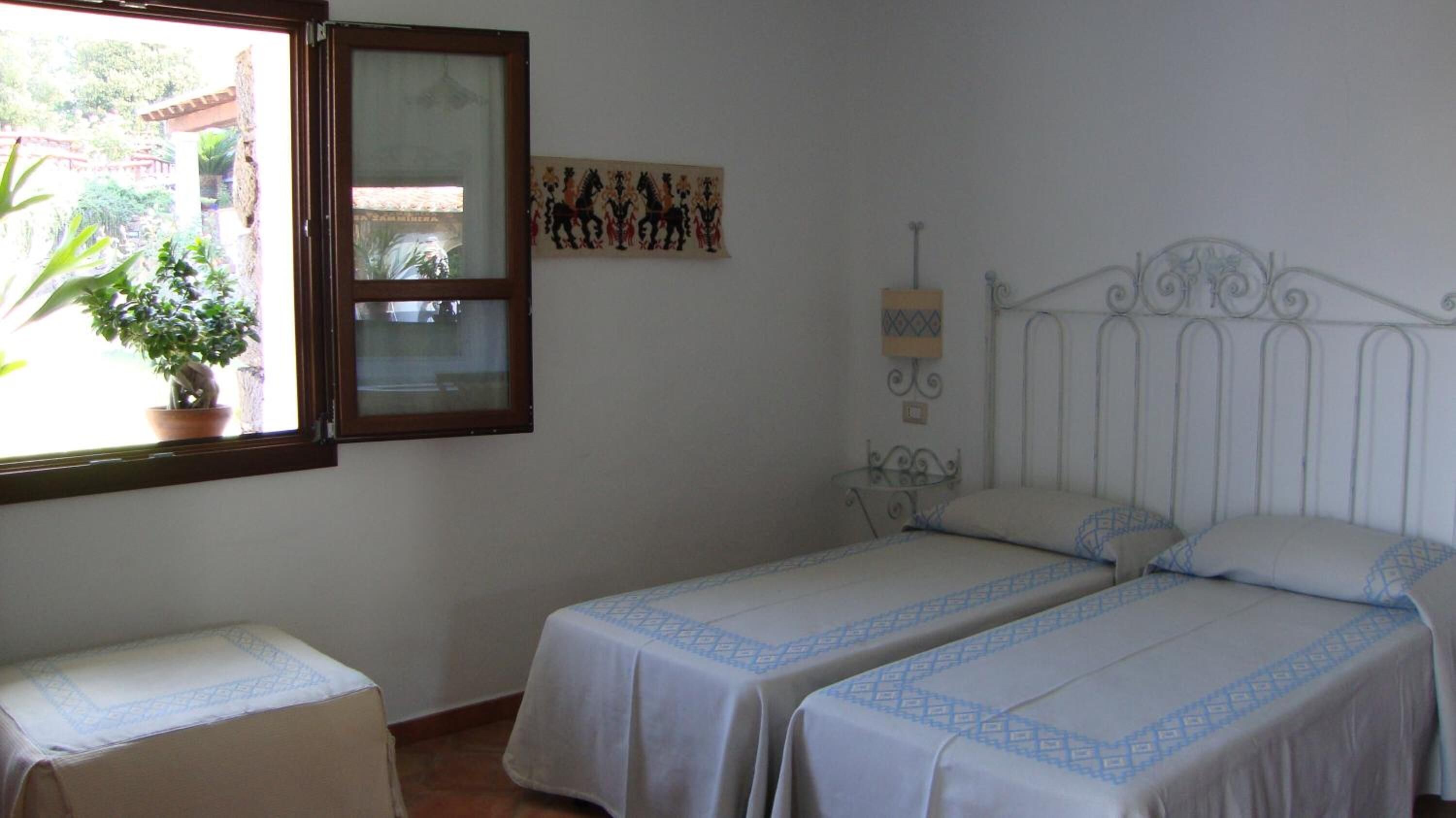 Un dormitorio con dos camas, una ventana con vista a follaje, una planta en maceta y una obra de arte colgada en la pared.