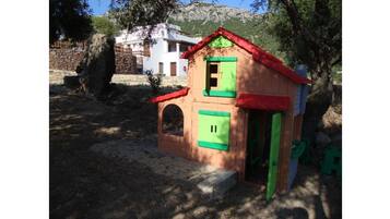 A small, colorful playhouse with a red roof and green shutters, situated in a natural setting with trees and a stone wall in the background.