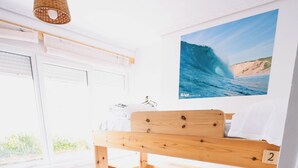 A wooden bunk bed with white bedding, a window with blinds, a wall-mounted picture of a wave, and a wicker hanging light fixture. - Latas Surf House (Ribamontan al Mar)