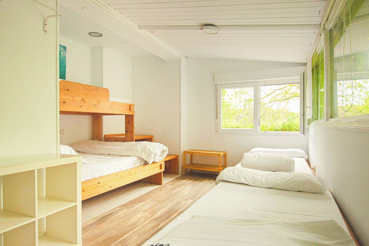 A room with two wooden bunk beds, a small wooden nightstand, and a window with a view of greenery.