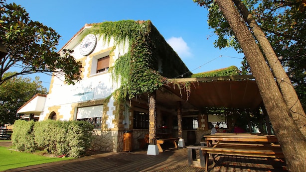 A building with ivy-covered walls, a clock, and a wooden deck with benches. - Latas Surf House (Ribamontan al Mar)