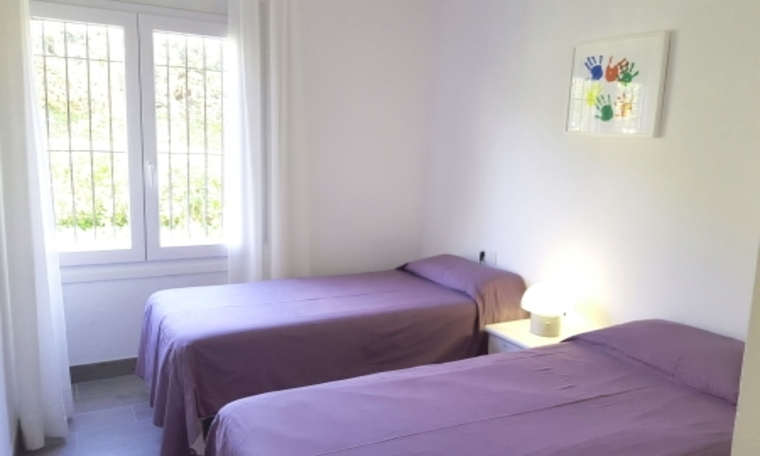 A room with two beds, a window with bars, a bedside table with a lamp, and a wall-mounted artwork.