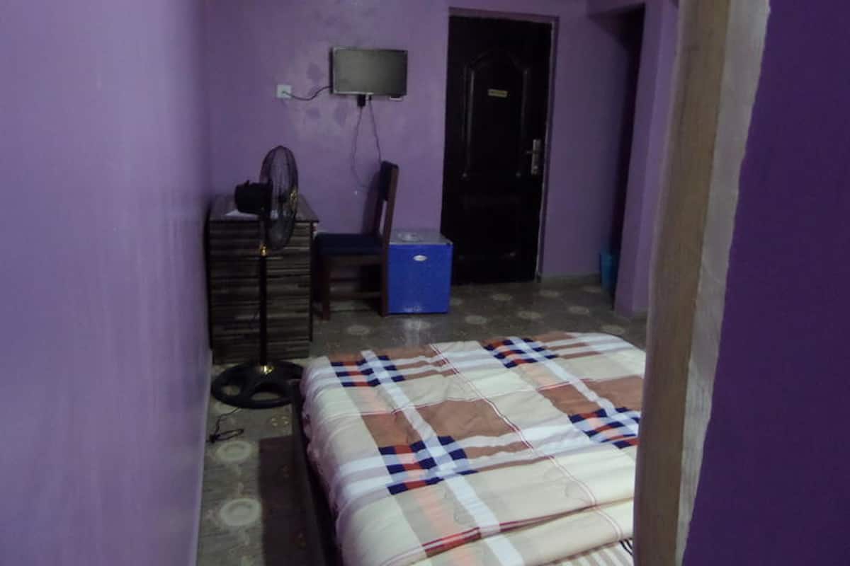 A small, dimly lit room with purple walls, a bed with a checkered blanket, a desk with a fan, a blue cooler, and a closed door.