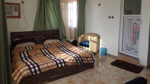 A bedroom with a bed, a chair, a door, and a window with curtains. - X Garden Hotel & Suites (Lagos)