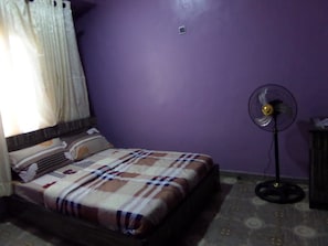 A room with a bed, a standing fan, and a window with curtains.