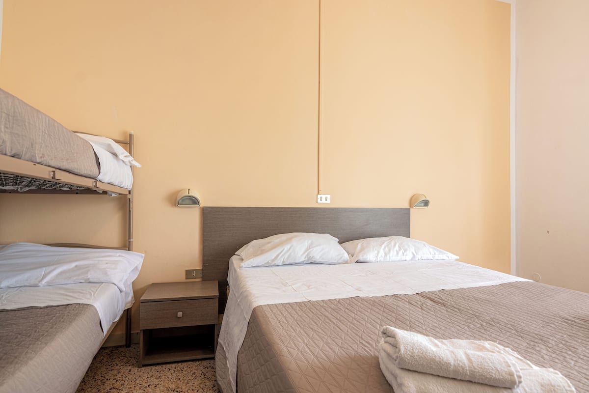 A hotel room with a bunk bed, a single bed, a nightstand, and a wall-mounted lamp.