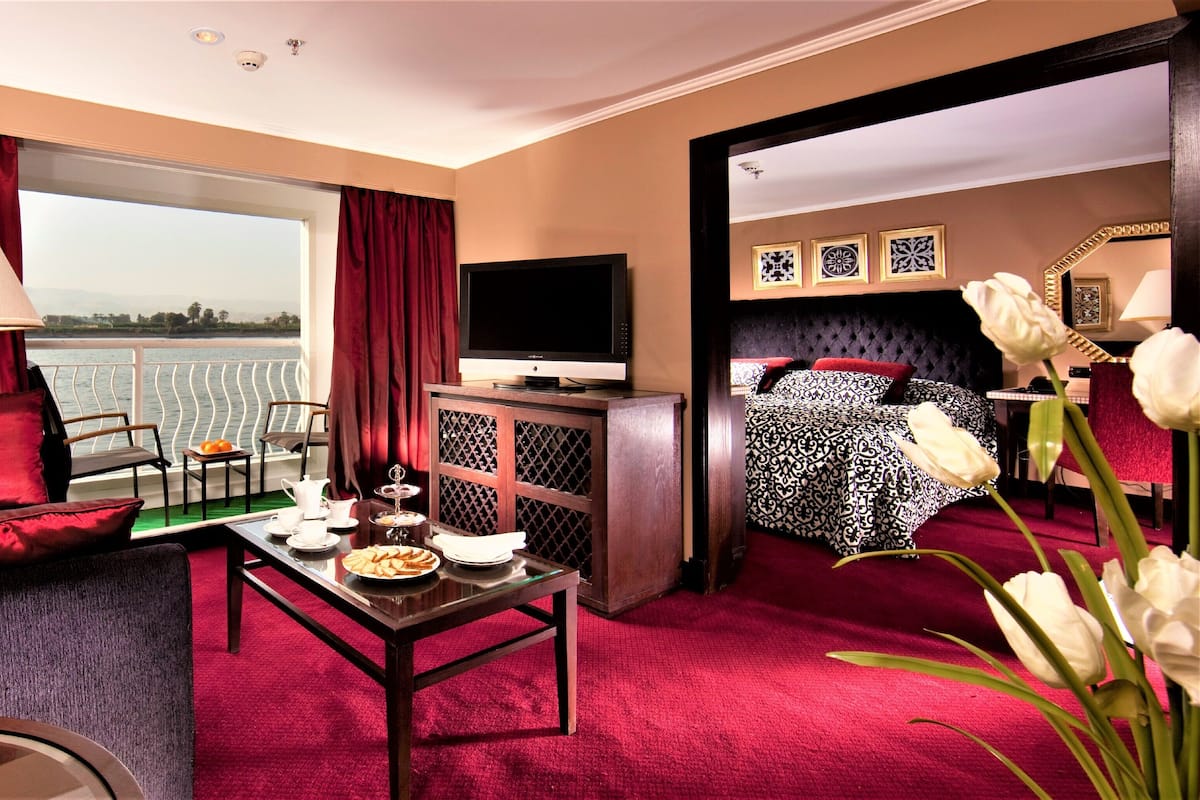 A hotel room with a large window, a television, a dining table with breakfast items, a bed with patterned bedding, and a red carpet.