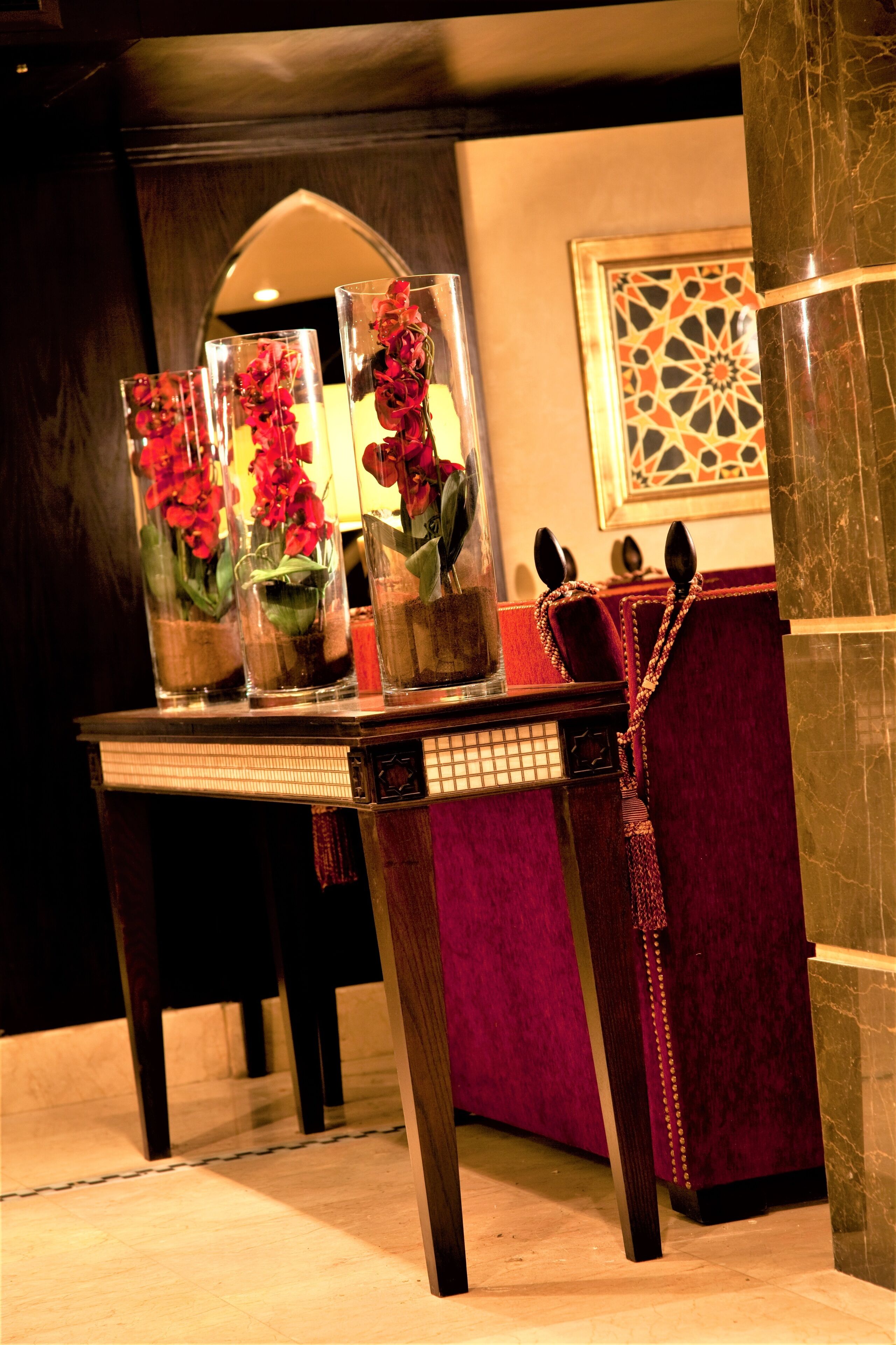 A table with three glass vases containing red flowers, a red upholstered chair, and a patterned wall decoration.