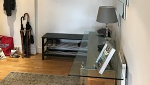Un salon moderne avec une table basse en verre, un canapé et une lampe. On y trouve un porte-manteau avec une veste et un sac, ainsi qu’une petite table avec une lampe.