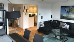 Un salon moderne avec une table basse en verre, des canapés noirs et un coin repas agrémenté d’une lampe noire.