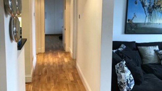 Un couloir avec un plancher en bois, un canapé noir orné de coussins à motifs et un tableau accroché au mur.
