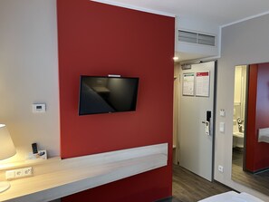 Habitación de hotel con una televisión montada en una pared roja, una mesita de noche con una lámpara y una puerta que da a un baño.