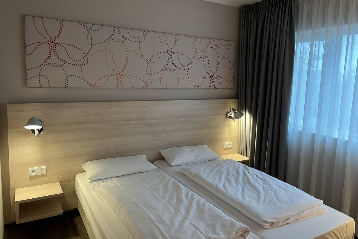A hotel room with a large bed, two bedside lamps, a wall art piece, and a window with curtains.