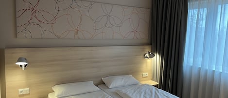 A hotel room with a large bed, two bedside lamps, a wall art piece, and a window with curtains.