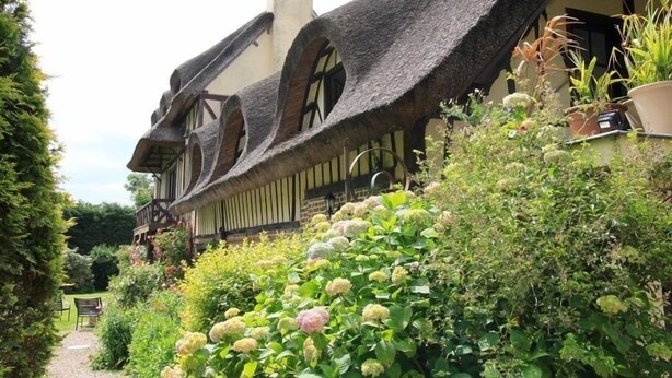 A traditional half-timbered house with a steeply pitched roof, surrounded by lush greenery and a gravel path leading to a bench. - Les Hauts d'Etretat (Bordeaux-Saint-Clair)