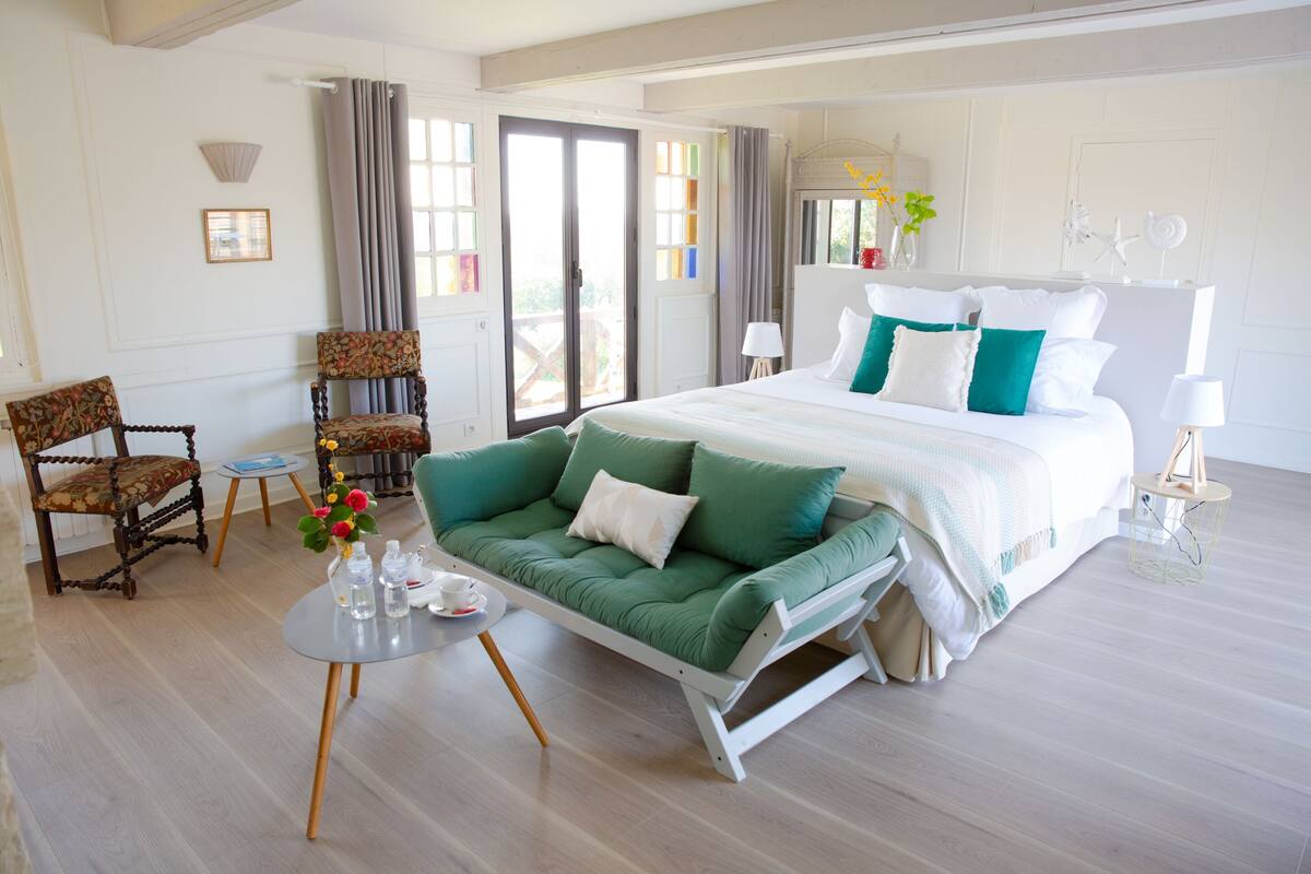 A bedroom with a bed, a green sofa, a chair, a small table with a vase of flowers, and a window with curtains.