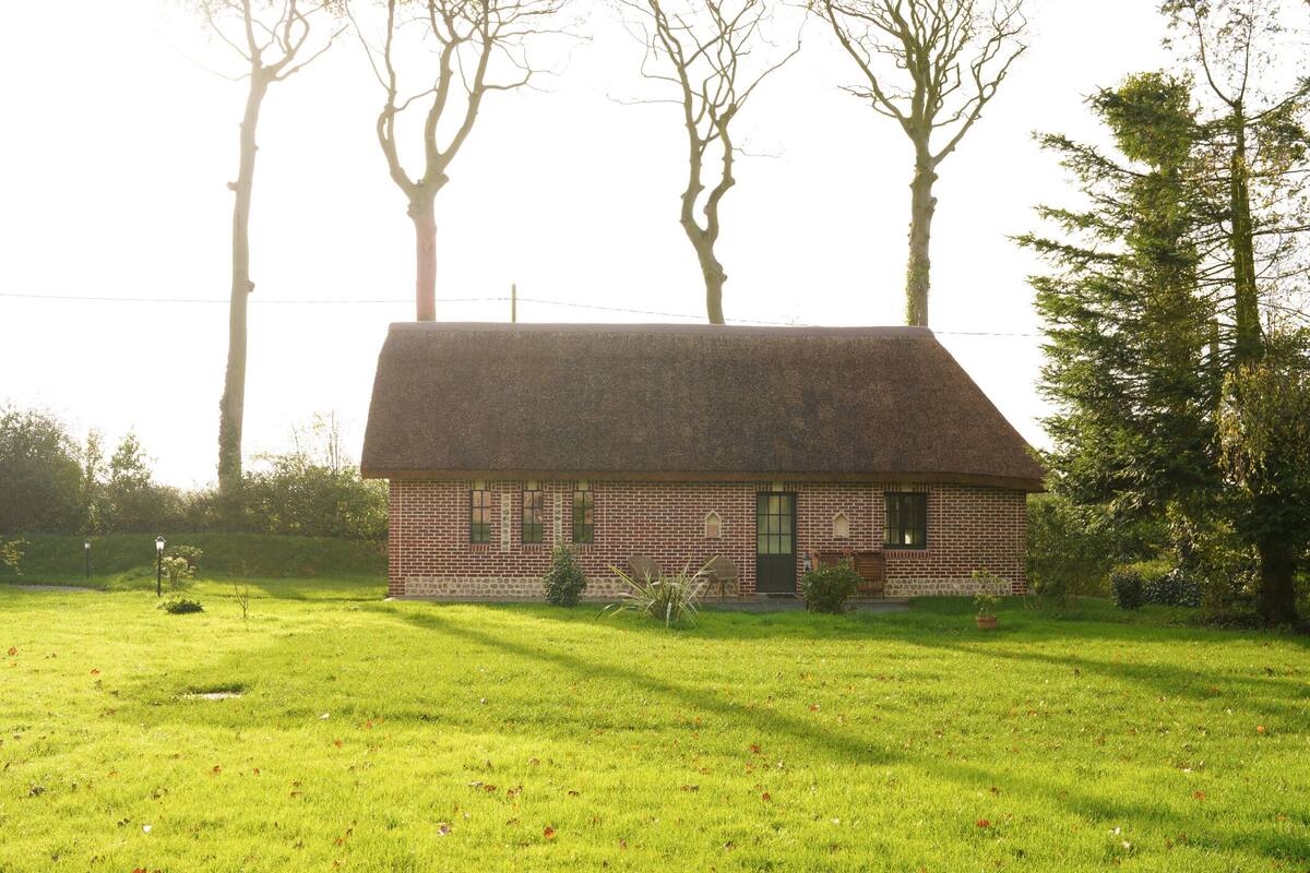 A brick house with a thatched roof in a grassy area, surrounded by trees.