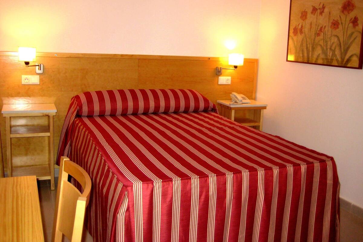 Habitación de hotel con cama, mesita de noche, silla, escritorio y teléfono instalado en la pared.