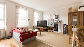 A cozy living room with a sofa, a television, a fireplace, and a dining area with wicker chairs. - Eson2 - The Kensington Gwendwr Road Lodge (London)